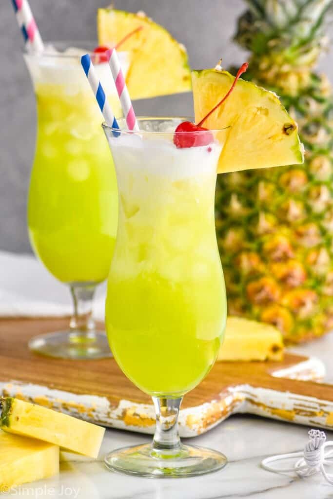 a hurricane glass filled with a midori splice garnished with a cherry and a pineapple wedge, some cream floating on top