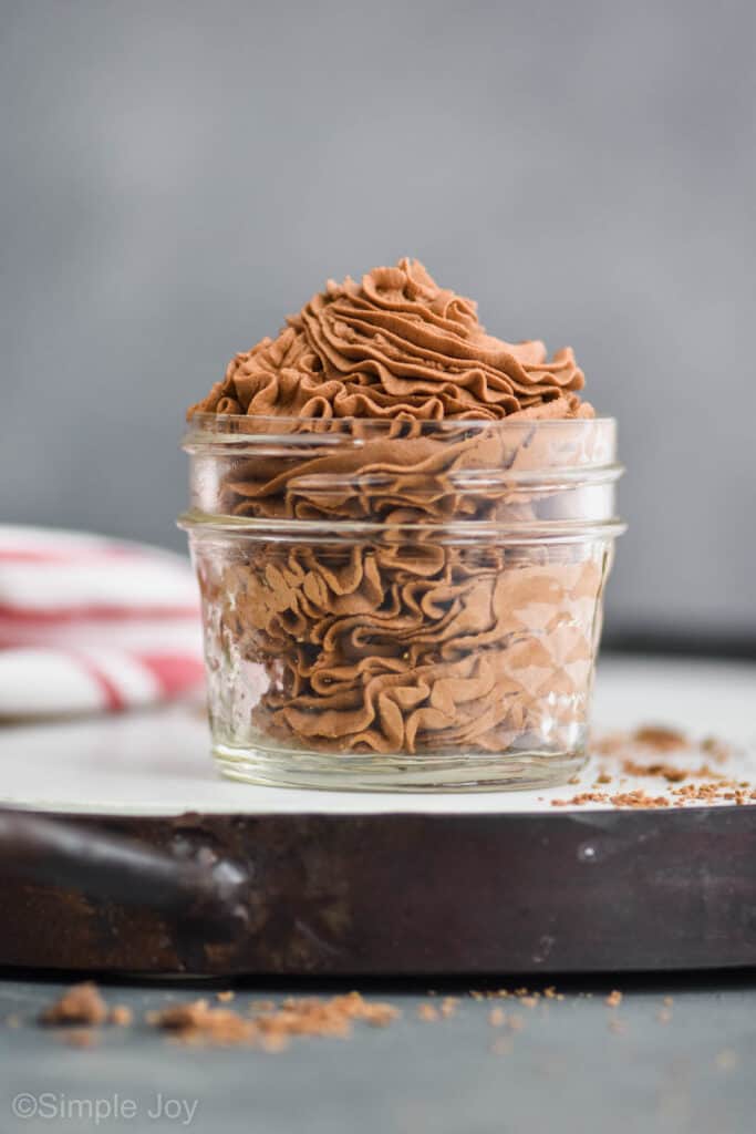 homemade chocolate whipped cream that has been piped into a small mason jar and is sitting on a white tray with chocolate shavings around it