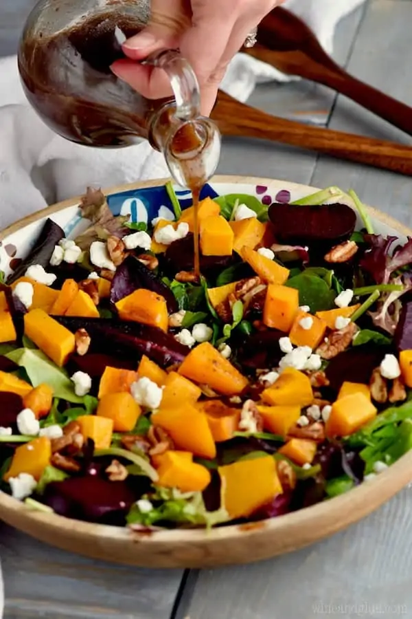 A close up photo of Roasted Beet Salad with cubbed butternut squash, pecans, goat cheese, mixed greens, roasted beats, and balsamic vinegar being poured. 