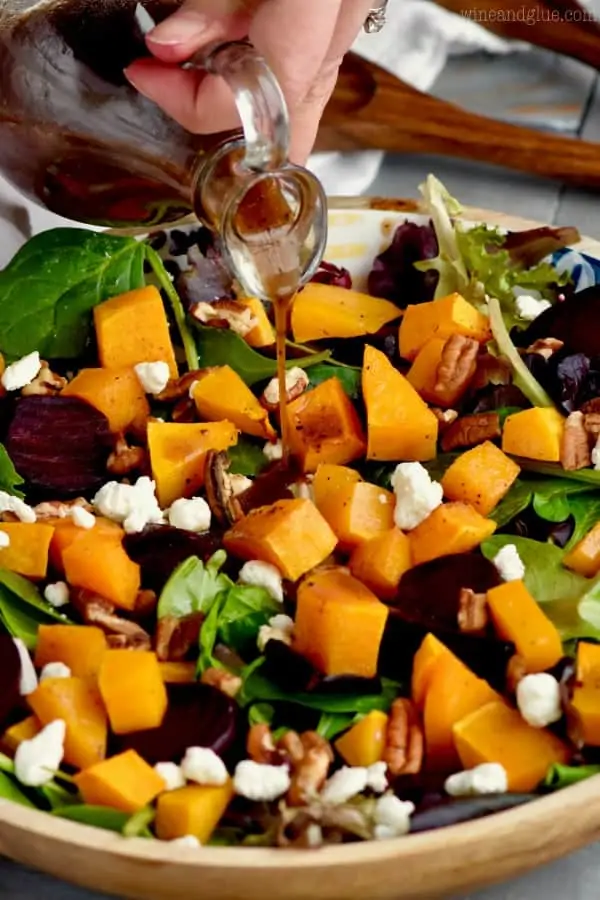 A close up photo of Roasted Beet Salad with cubbed butternut squash, pecans, goat cheese, mixed greens, roasted beats, and balsamic vinegar being poured.  