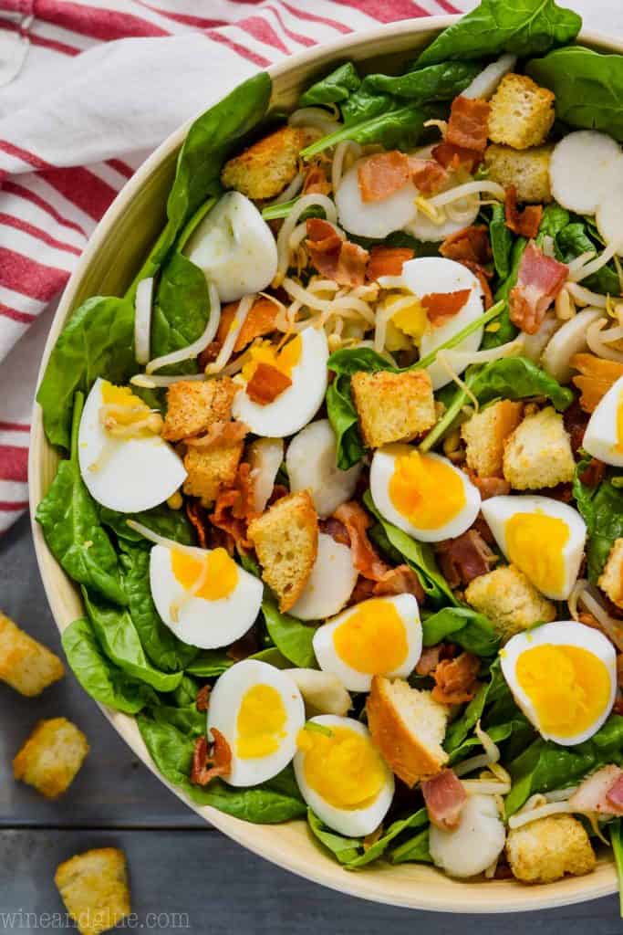 overhead view of spinach bacon salad with hard boiled eggs, croutons, water chestnuts in a white bowl