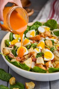 woman's handing pouring a mason jar full of red spinach salad dressing onto a spinach bacon salad with hard boiled eggs and croutons in a white bowl