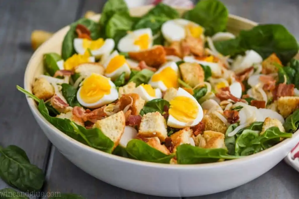 white bowl filled with spinach salad with bacon, hard boiled eggs, and croutons on a blue wood board