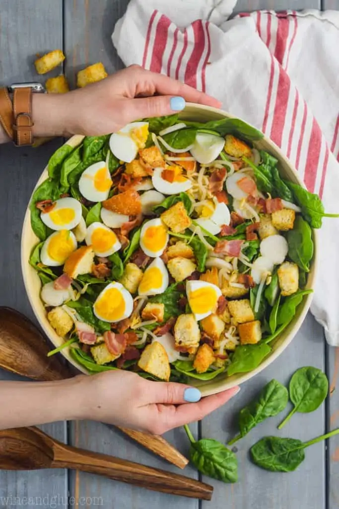 two hands holding a white salad bowl filled with the best spinach salad topped with sliced boiled eggs, bacon, and cheese. 