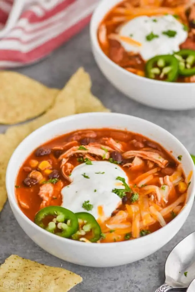 white bowl on a gray surface with sour cream, shredded cheese and jalapeños garnishing chicken taco soup, another bowl int he background