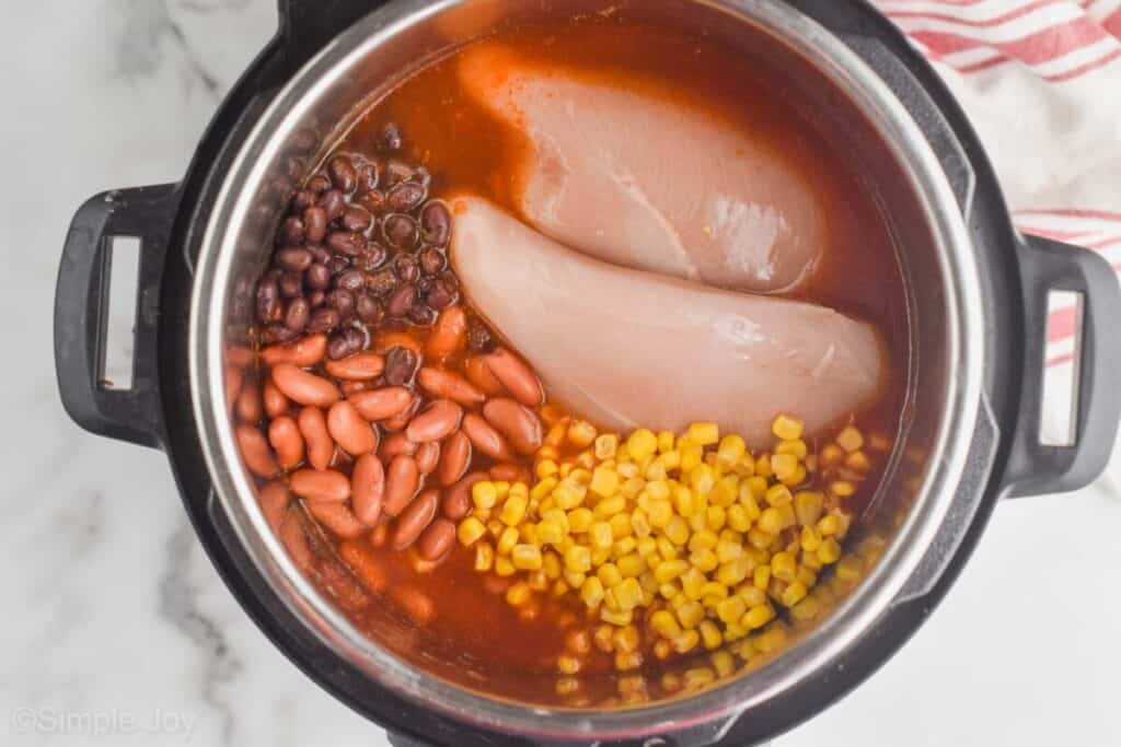 overhead view of an instant pot with all the ingredients for taco soup in it: black beans, kidney beans, corn, chicken, crushed tomatoes, taco seasoning, and chicken stock