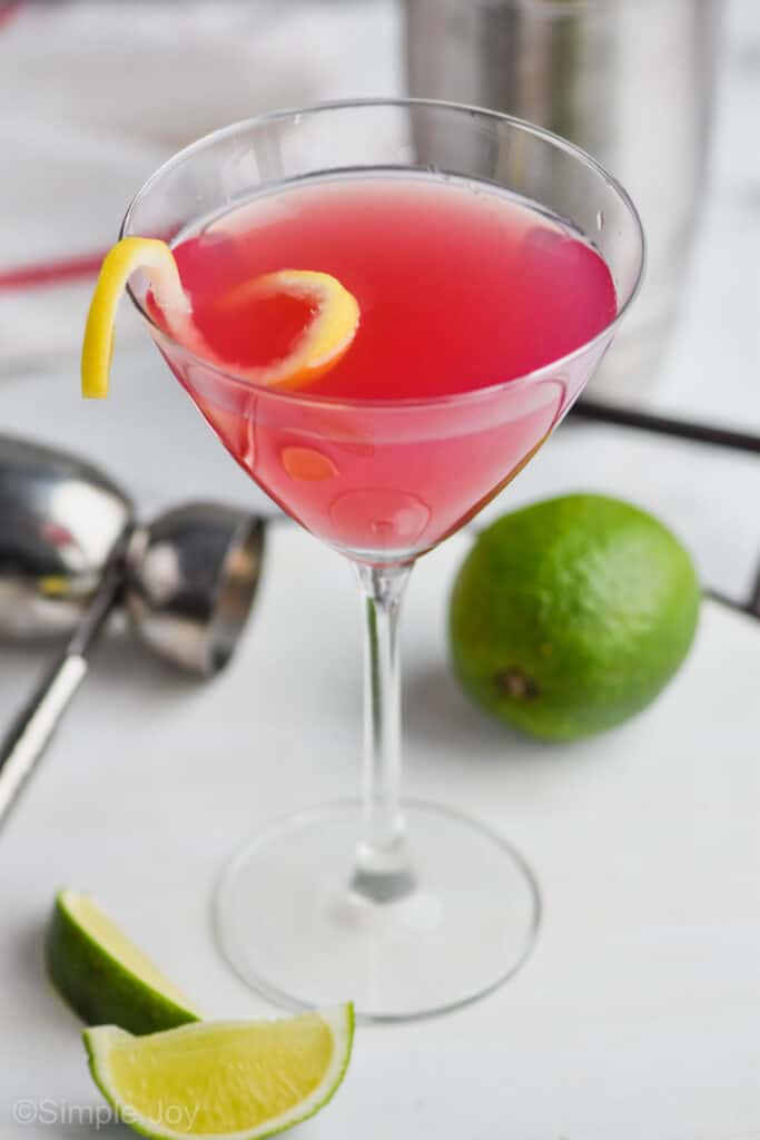 overhead view of a martini glass filled with pink cosmopolitan drink recipe with a lemon curl on a white tray with a silver jigger and a lime next to it