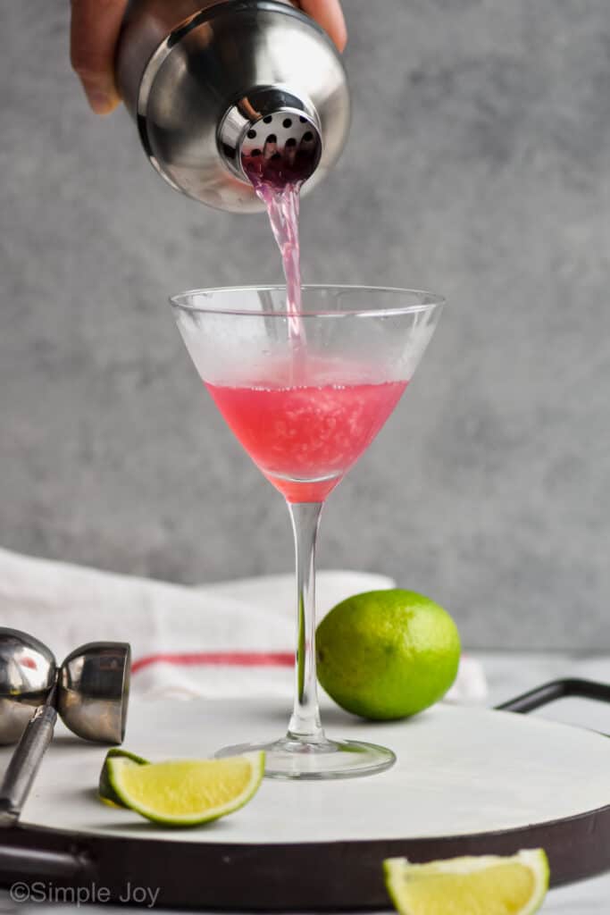 a silver cocktail shaker pouring a pink cosmopolitan recipe into a frosted martini glass that is sitting on a white tray with a lime next to it and a metal jigger on the other side