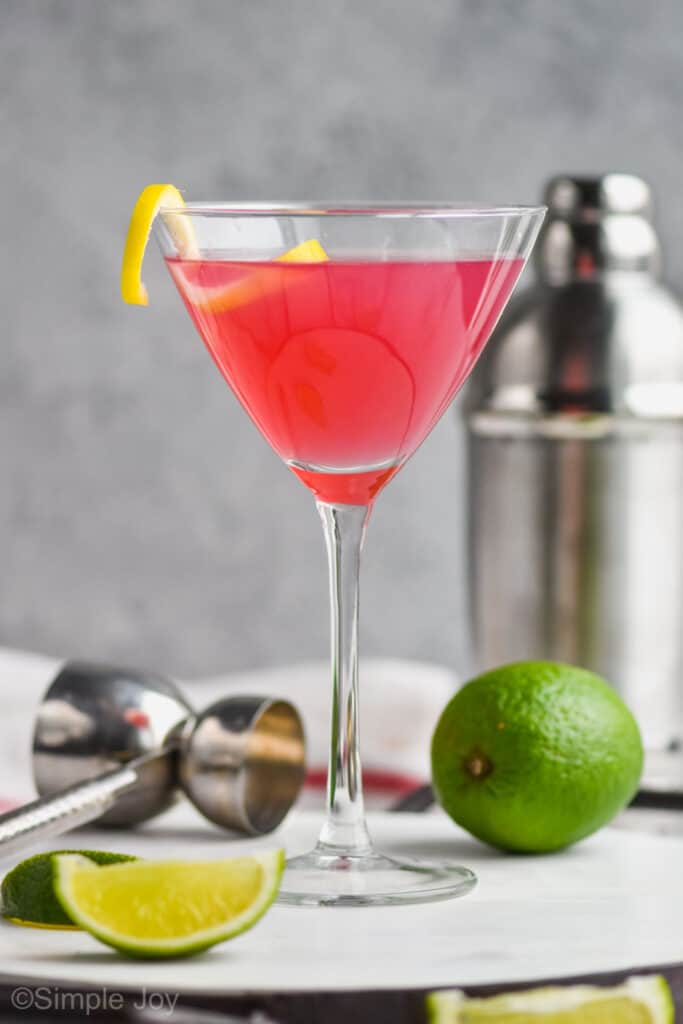 martini glass on a tray with a metal jigger, and metal cocktail shaker int he background, filled with cosmopolitan drink garnished with a lemon curl