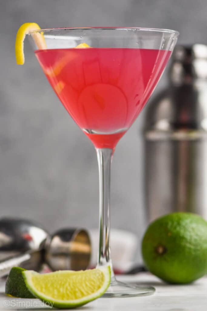 close up of a cosmopolitan recipe in a martini glass, garnished with a lemon curl, with a silver cocktail shaker in the background
