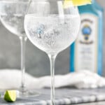 Two glasses of the perfect gin and tonic recipe, each garnished with a lime wedge. Bottle of gin sits in the background and lime wedges sit on the counter. - 8
