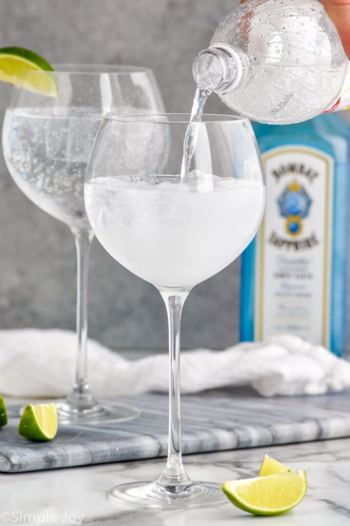 Man's hand pouring gin for the perfect gin and tonic recipe into glass with ice. Bottle of gin sits in the background, and lime wedges sit on the counter.