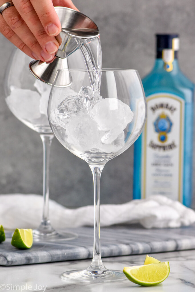Man's hand pouring gin for the perfect gin and tonic recipe into glass with ice. Bottle of gin sits in the background, and lime wedges sit on the counter.