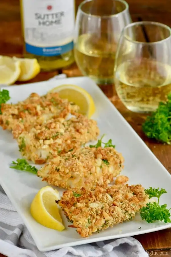  five pieces of oven fried fish on a platter with a slice of lemon and a glass of white wine