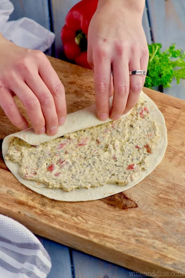 On a wooden cutting board, a woman is rolling the tortilla with Pest Cream Cheese on it.