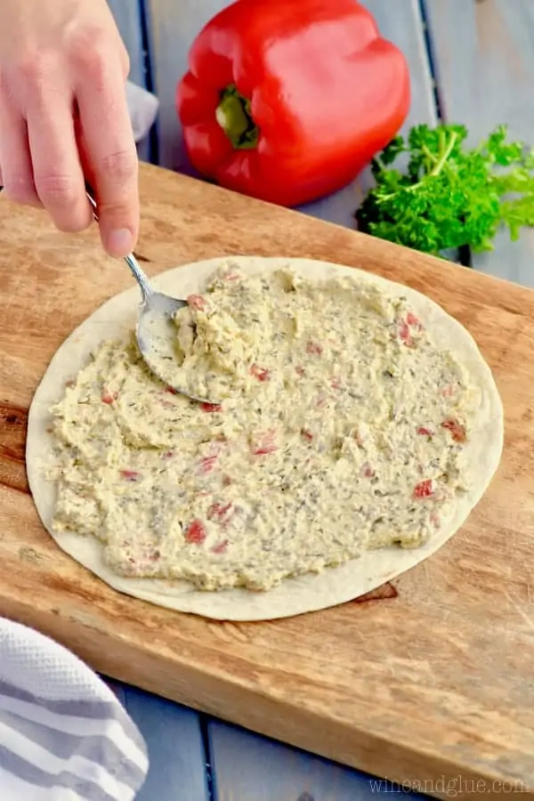 On a wooden cutting board, a woman with a spoon is spreading the Pesto Cream Cheese onto the tortilla 
