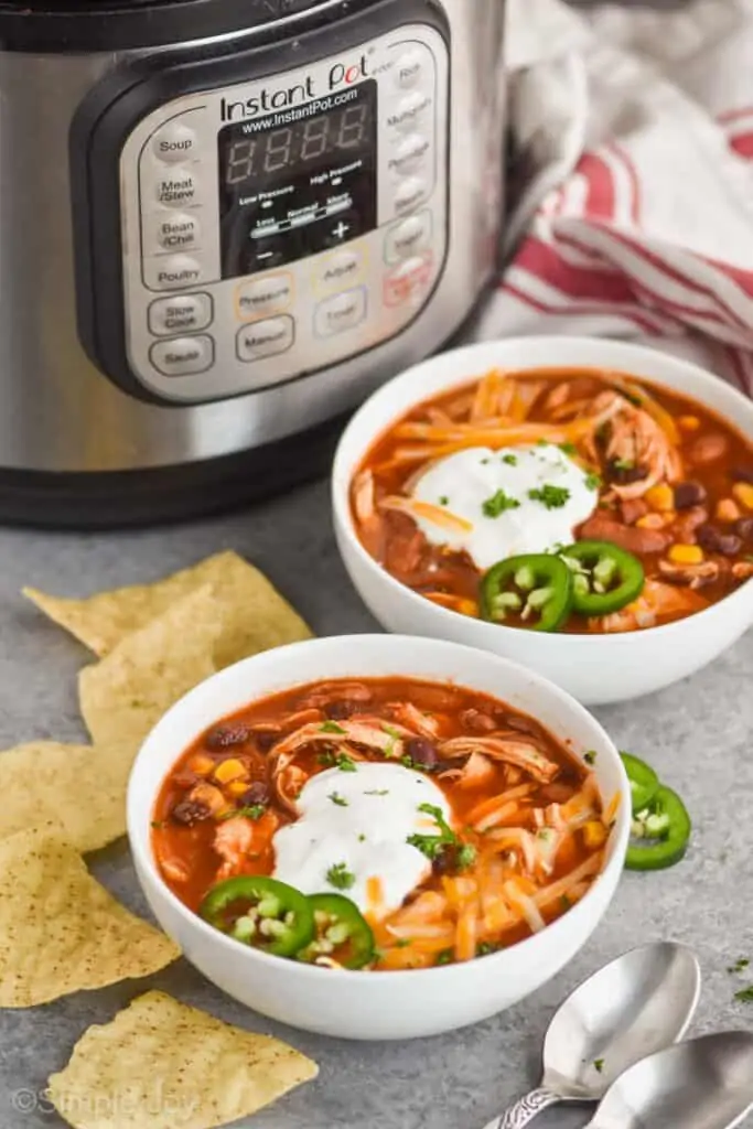 two white bowls full of chicken taco soup topped with sour cream and cheese  with tortilla chips around them and an instant pot int he background