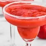 two margarita glasses rimmed with sugar and filled with strawberry margaritas surrounded by strawberries and limes