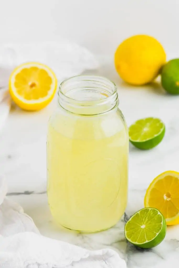 clear mason jar full of homemade sweet and sour mix with limes and lemons around it on marble counter top