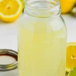 clear mason jar full of sweet and sour mix with limes and lemons around it on marble counter top