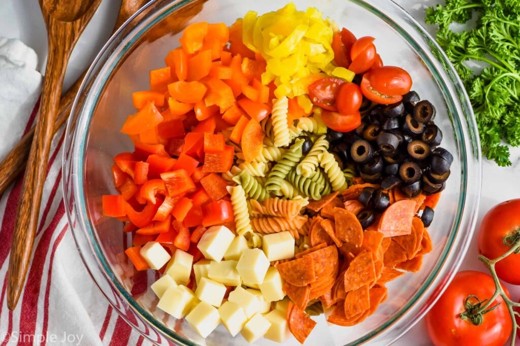 overhead of a bowl of italian pasta salad ingredients, with pasta, orange and red bell pepper, banana peppers, cherry tomatoes, olives, pepperoni, and cheese cubes