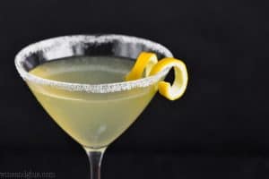a land scape photo of the top half of a martini glass rimmed with sugar, full of a lemon drop martini recipe, and garnished with a lemon curl on a black background
