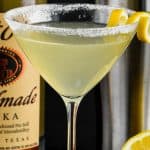 a lemon drop martini recipe in a martin glass, rimmed with sugar, garnished with a lemon curl and a bottle of titos and a martini shaker in the background