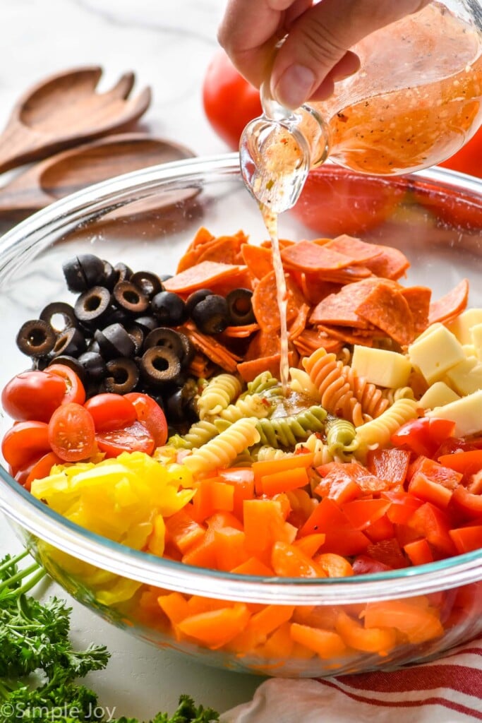 pouring italian dressing over a bowl of ingredients for Italian pasta salad