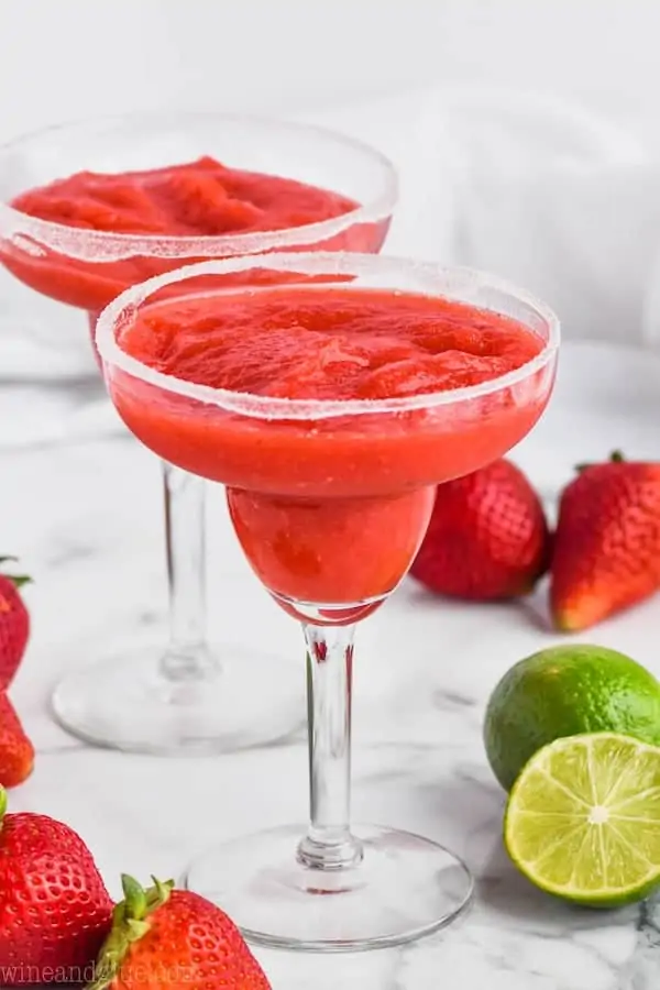 two margarita glasses rimmed with sugar and filled with strawberry margaritas surrounded by strawberries and limes
