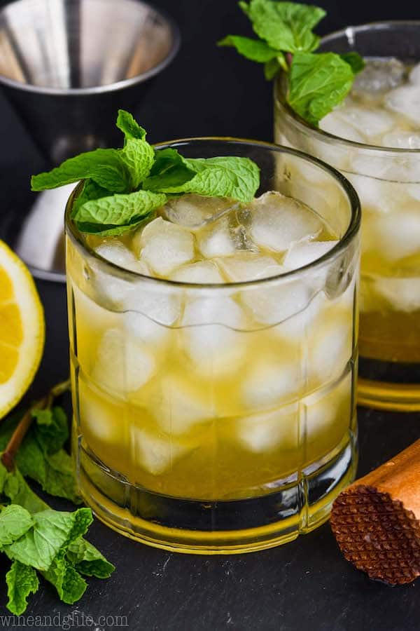 a small tumbler filled with ice and whiskey smash recipe, garnished with fresh mint with a lemon, another cocktail, and meddler in the background