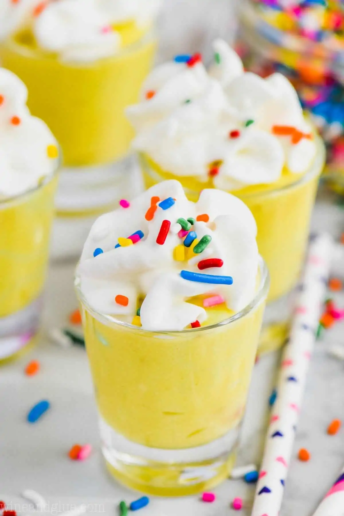 shot glass filled with birthday cake pudding shots, topped with whipped cream and rainbow sprinkles, three other filled shot glasses in the background