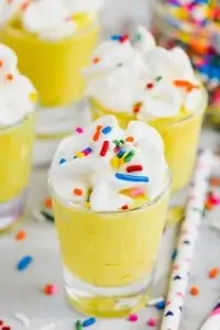 shot glass filled with birthday cake pudding shots, topped with whipped cream and rainbow sprinkles, three other filled shot glasses in the background