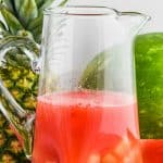 a tall pitcher of watermelon rum punch with a pineapple and a watermelon in the background