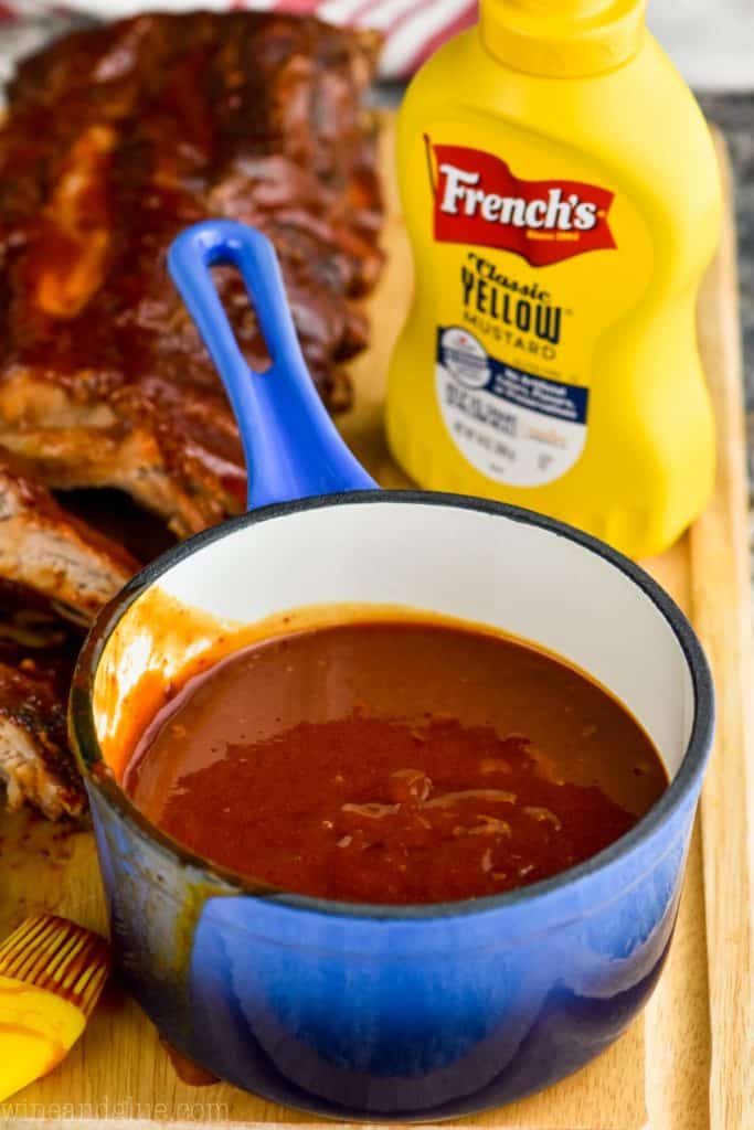 small sauce pan of barbecue sauce with ribs in the background and yellow mustard. 