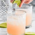 fingers squeezing a lime wedge into a tumbler half filled with ice and a paloma cocktail