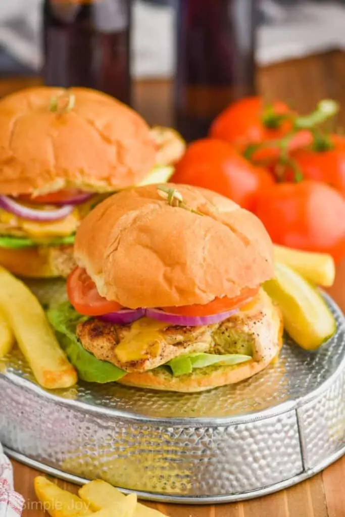 pulled back view of a two grilled chicken sandwiches on a turned over metal try with steak fries around them, sandwiches held together with a toothpick