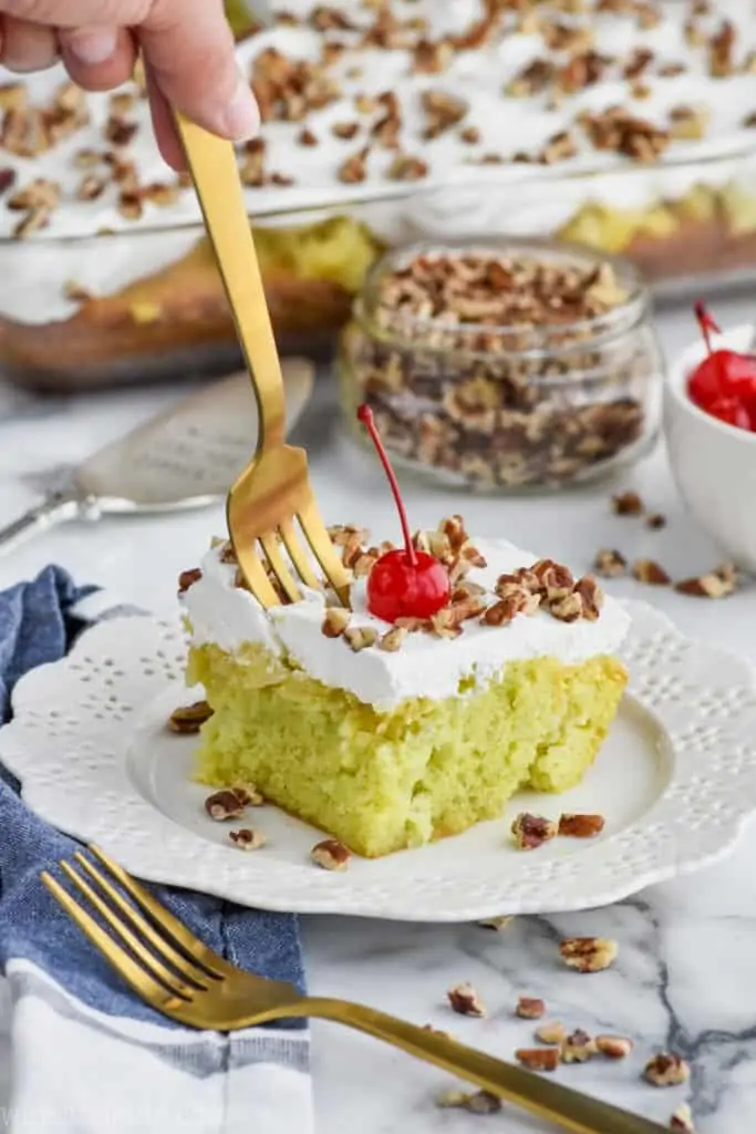 A piece of Watergate Cake topped with white frosting, cherries, and walnuts.