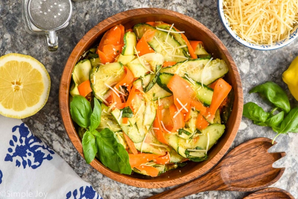 Overhead view of a bowl of Zucchini Salad. Salad tongs, half of a lemon, bowl of shredded cheese, salt, and basil beside bowl.