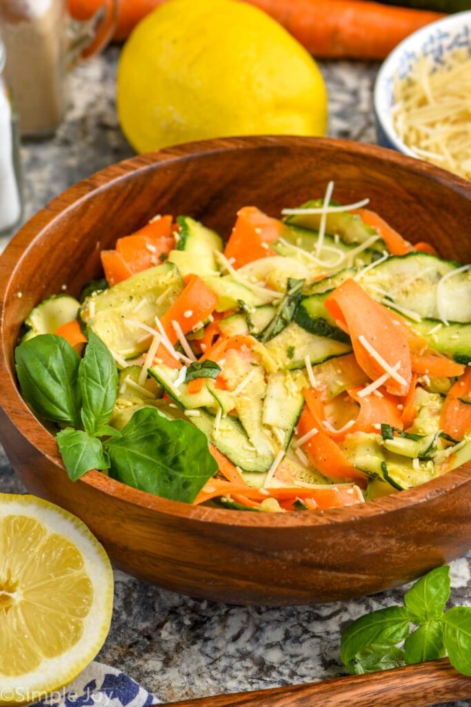 Side view of a bowl of Zucchini Salad. Lemons and bowl of shredded cheese beside bowl.