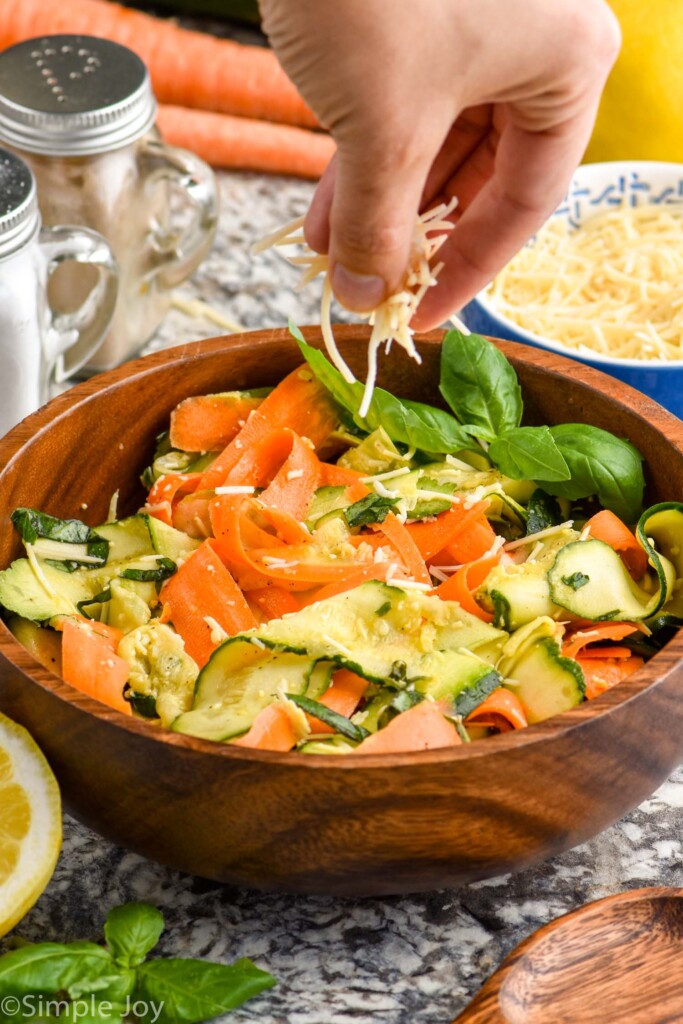 Side view of person's hand sprinkling shredded cheese on top of Zucchini Salad.