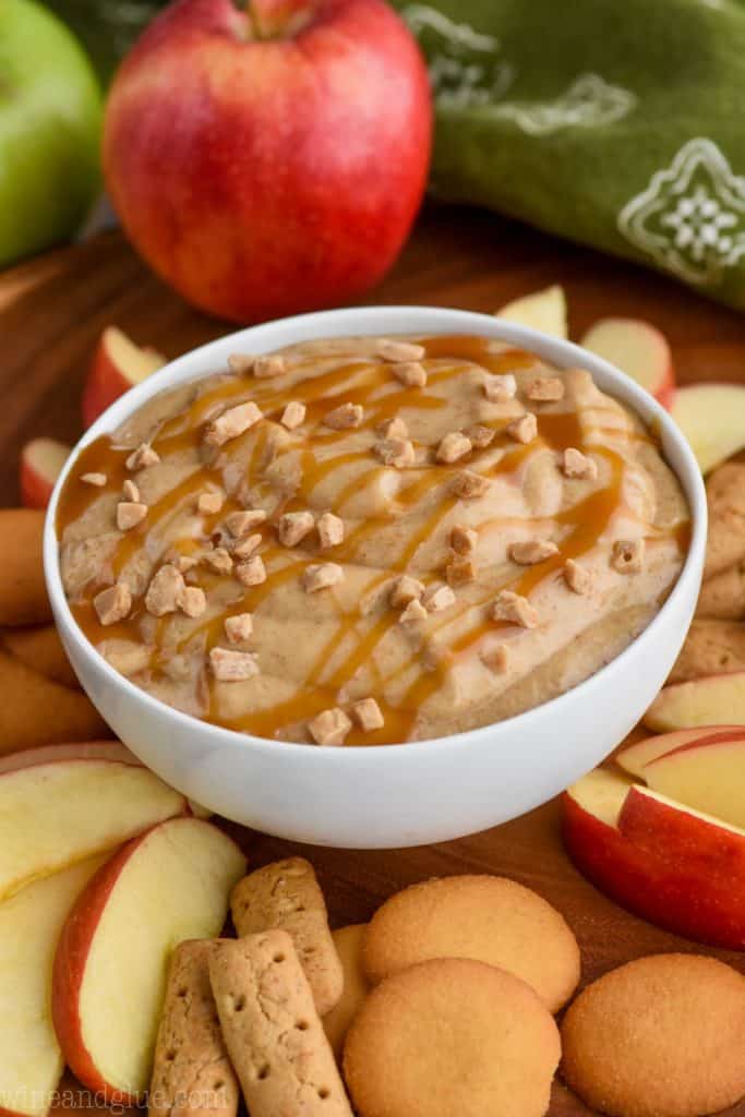 In a white bowl, the Caramel Apple Dip is topped with crushed toffee and caramel and surrounded with apples, graham crackers, and wafers cookies. 