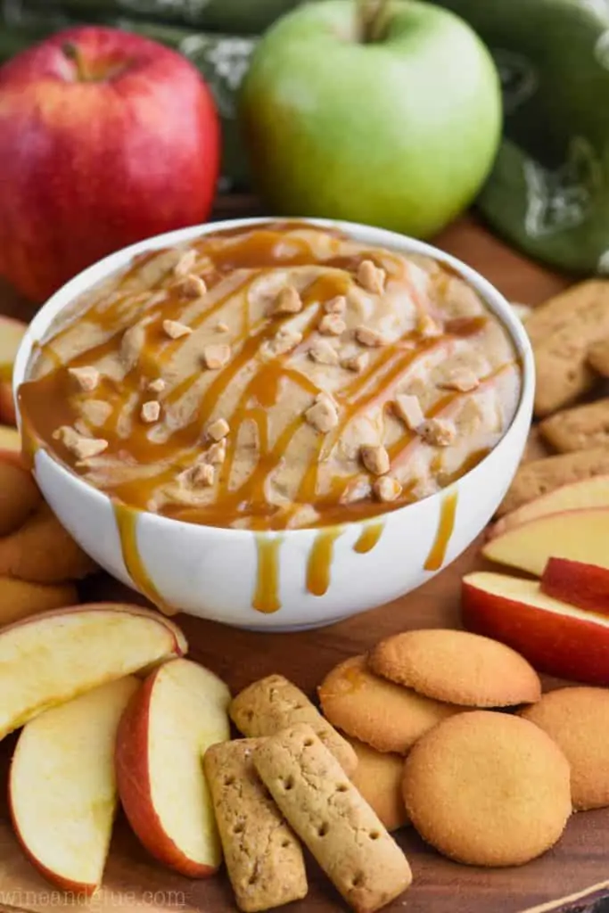 In a small white bowl, the Caramel Apple Dip sauce is topped with caramel and toffee bits and surrounded with apples, graham crackers, and wafers. 