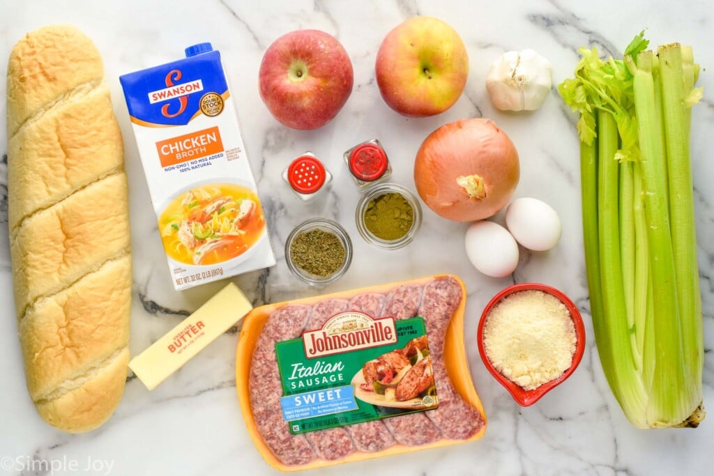 a picture of ingredients for homemade stuffing, loaf of French bread, chicken stock, butter, chicken stock, sausage, sage, thyme, salt and pepper, apples, onion, eggs, garlic, celery, and cheese