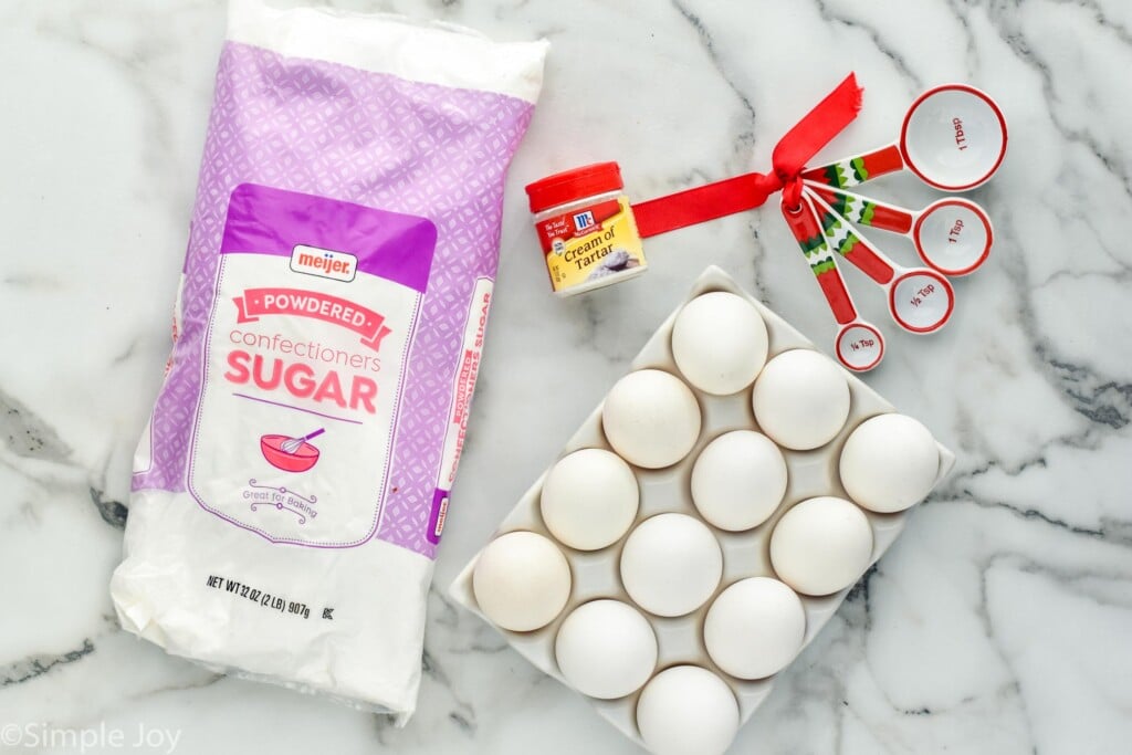 overhead photo of ingredients for gingerbread house icing: powdered sugar, eggs, cream of tartar with a Christmas themed measuring spoon set