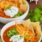 two white bowls filled with slow cooker chicken tortilla soup, garnished with sour cream, shredded cheese, cilantro, jalapeno slices, and tortilla chips