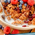 pinterest graphic of bowl of homemade granola with milk, a spoon, fresh raspberries and blueberries, says: "the best granola simplejoy.com"