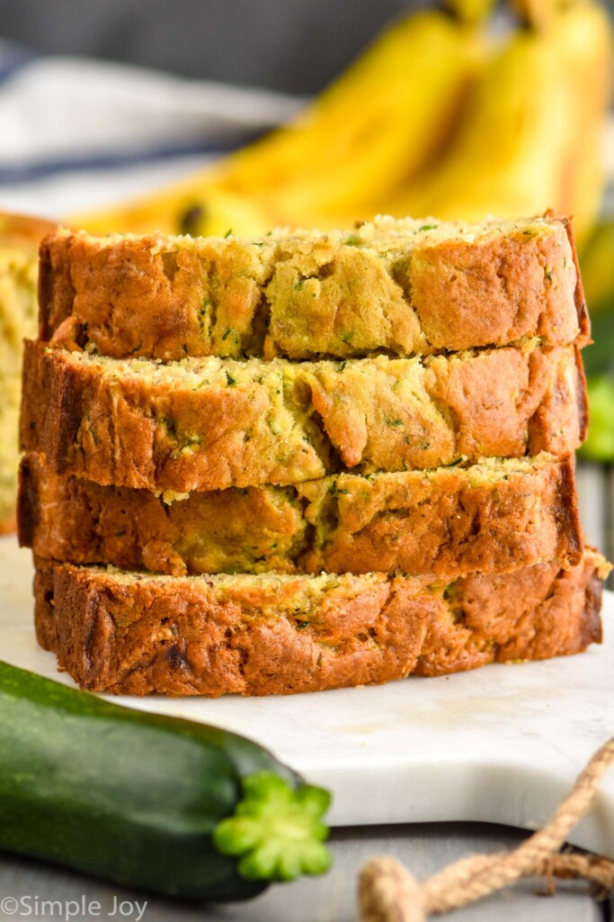 Close up photo of a stack of slices of Banana Zucchini Bread. Bananas and Zucchini sit beside bread.