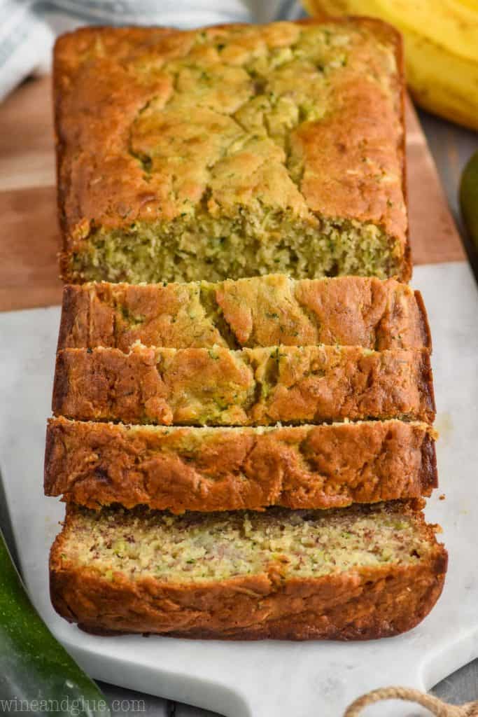 front view of zucchini banana bread recipe sliced on a marble and wood cutting board surrounded by two zucchini and a bunch of bananas on a gray wood board