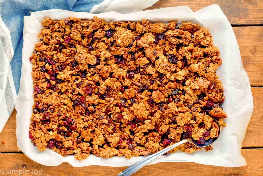 baking sheet covered parchment paper with homemade granola recipe and metal spoon from overhead on a wooden board
