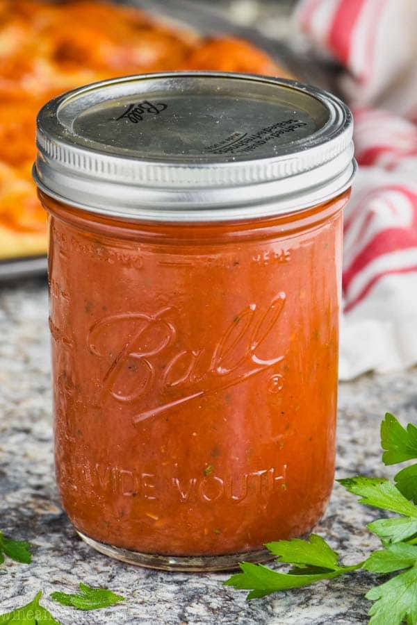 ball jar filled with homemade pizza sauce recipe surrounded by parsley leaves with pizza in the background